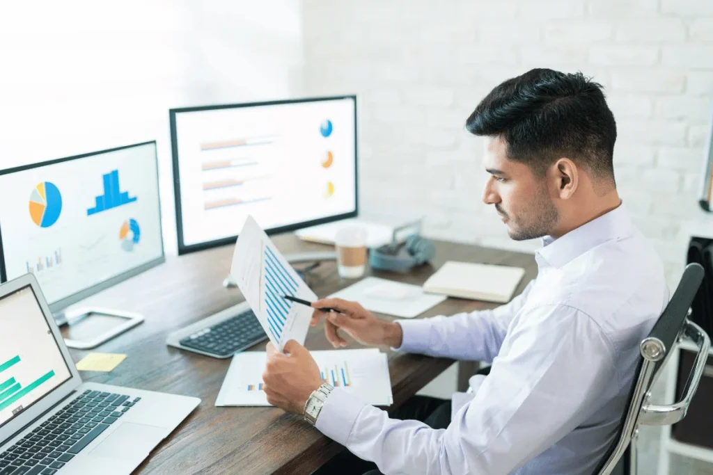 A man in a white shirt sits at a desk analyzing graphs and charts on paper and computer screens, conveying focus and analysis in a modern office.
