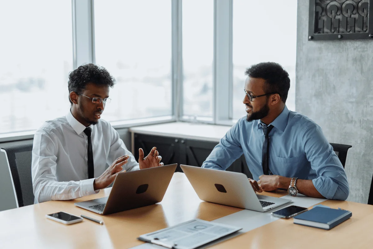 Two professionals are engaged in discussion at a meeting table, with laptops and a notebook visible, set against a large window backdrop.