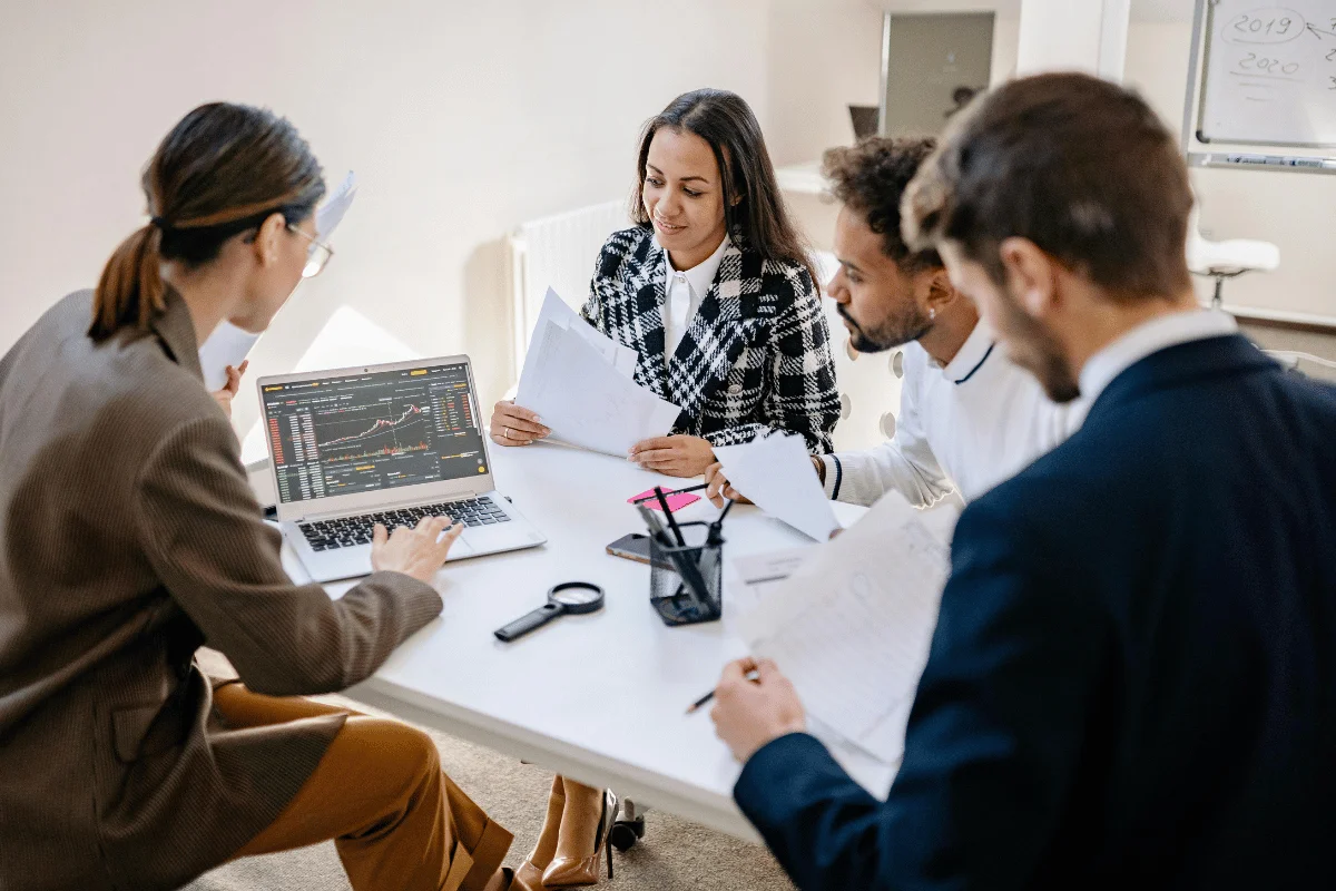 Four people in formal attire sit around a table, reviewing documents and analyzing a laptop displaying stock market data. 