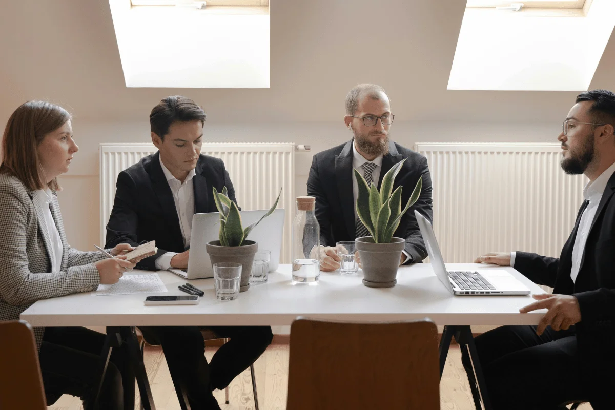Four people in business attire sit around a table with laptops and potted plants, engaged in a serious meeting under natural light from a window.