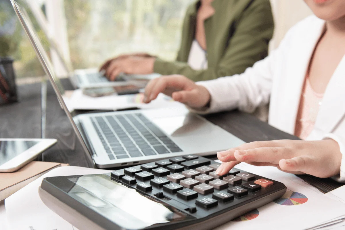 Two hands work on a calculator and laptop, surrounded by documents and a tablet, in a bright, collaborative workspace.