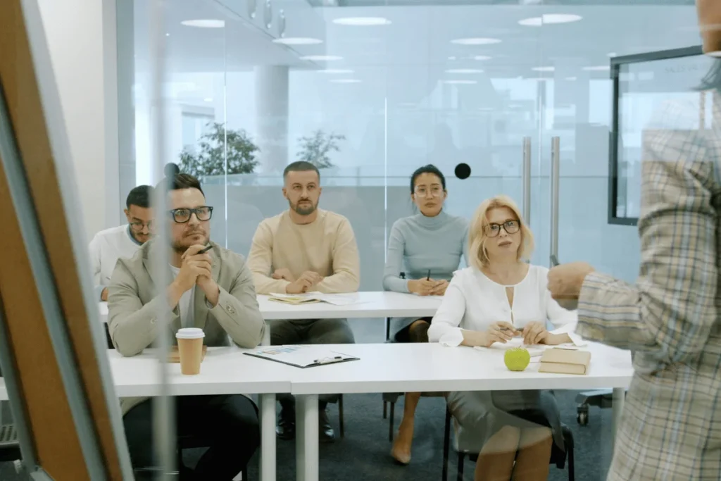 A group of people seated in a modern meeting room, attentively listening to a speaker while taking notes and sipping coffee.