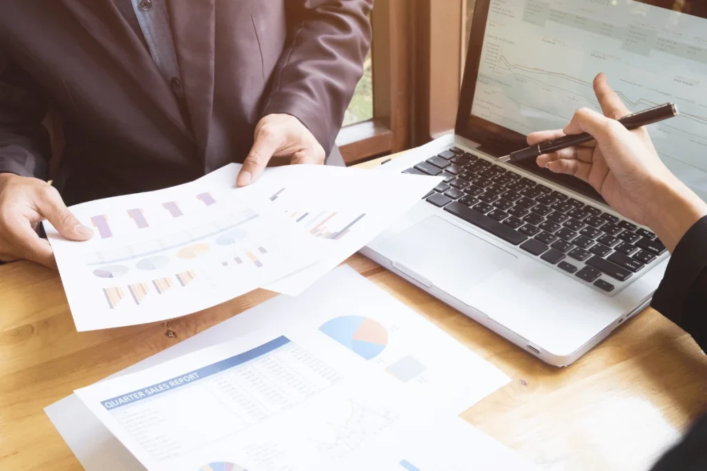 Two people review financial charts at a wooden table, with documents and a laptop displaying graphs. 
