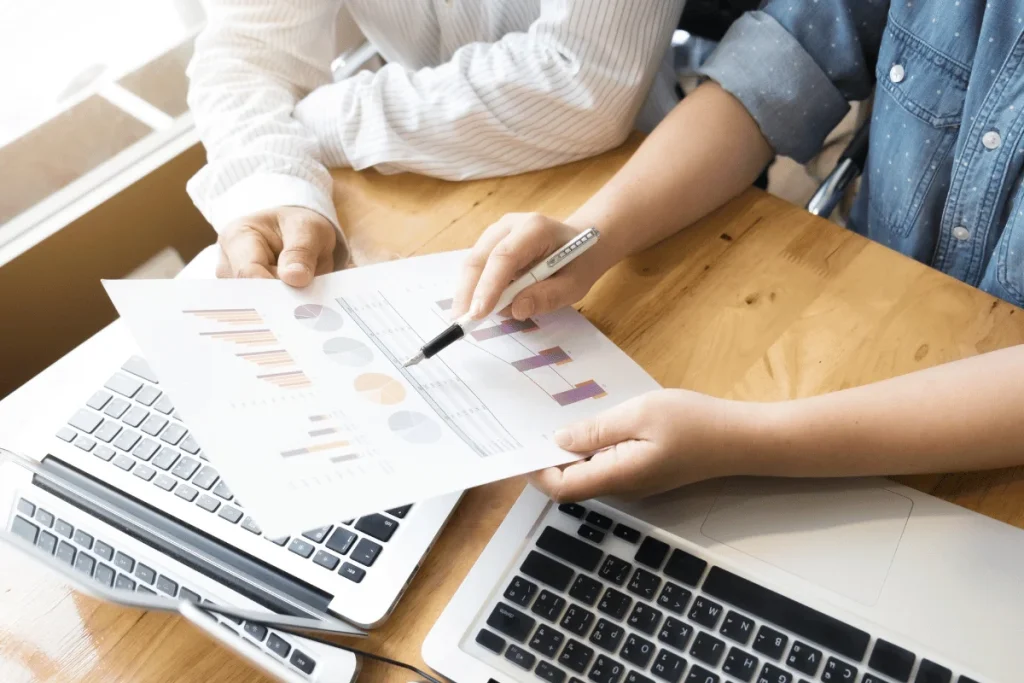 Two people reviewing a chart with bar and pie graphs on paper over laptops at a wooden table.