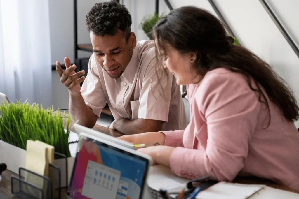 Two people are discussing at a desk with plants and a laptop, conveying collaboration.