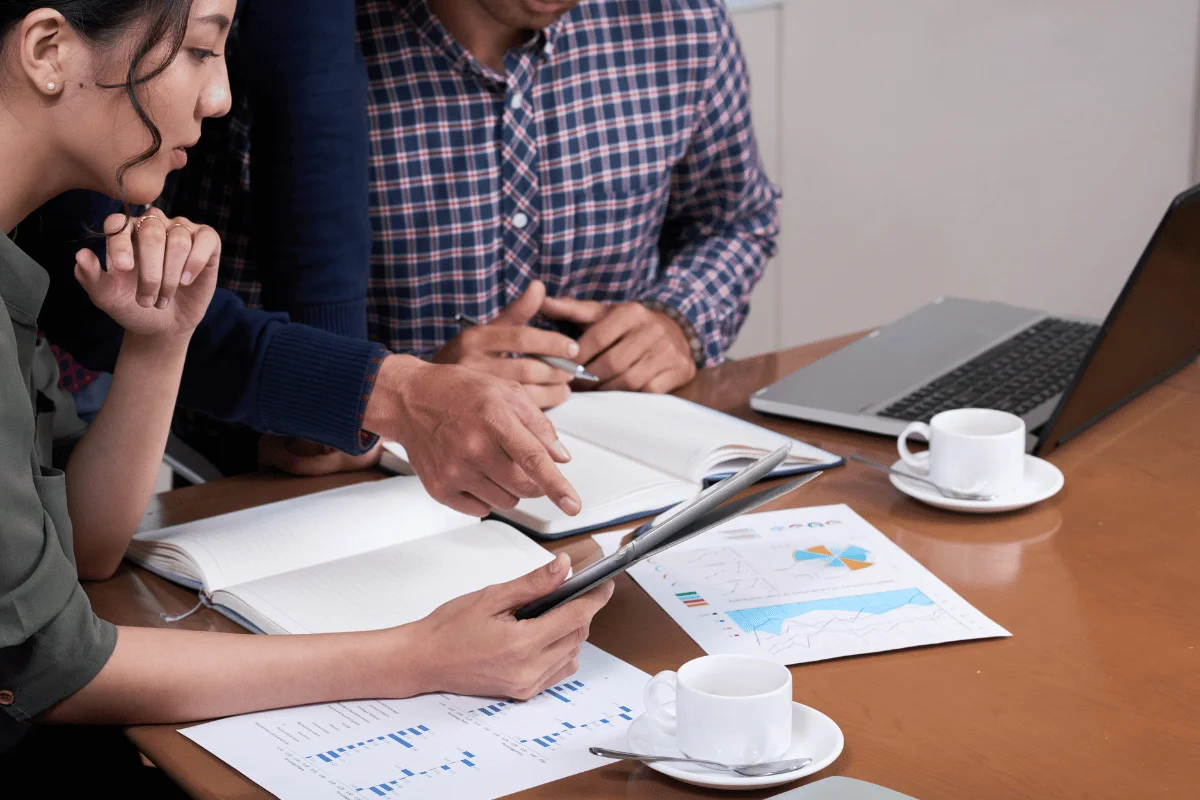 Three people are gathered around a table with charts, a laptop, and coffee cups, analyzing data. 