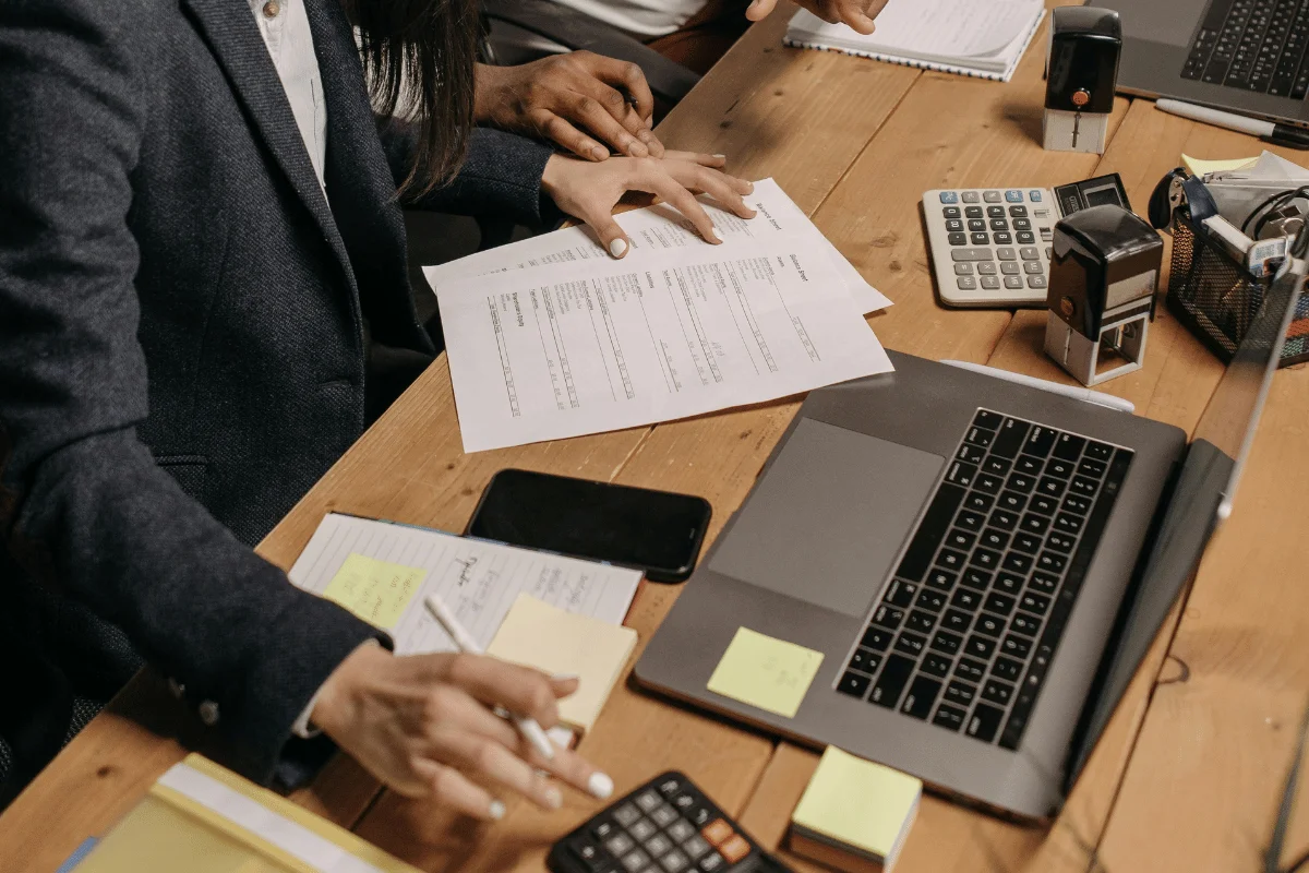Two coworkers review printed documents at a wooden desk with laptop and calculator.