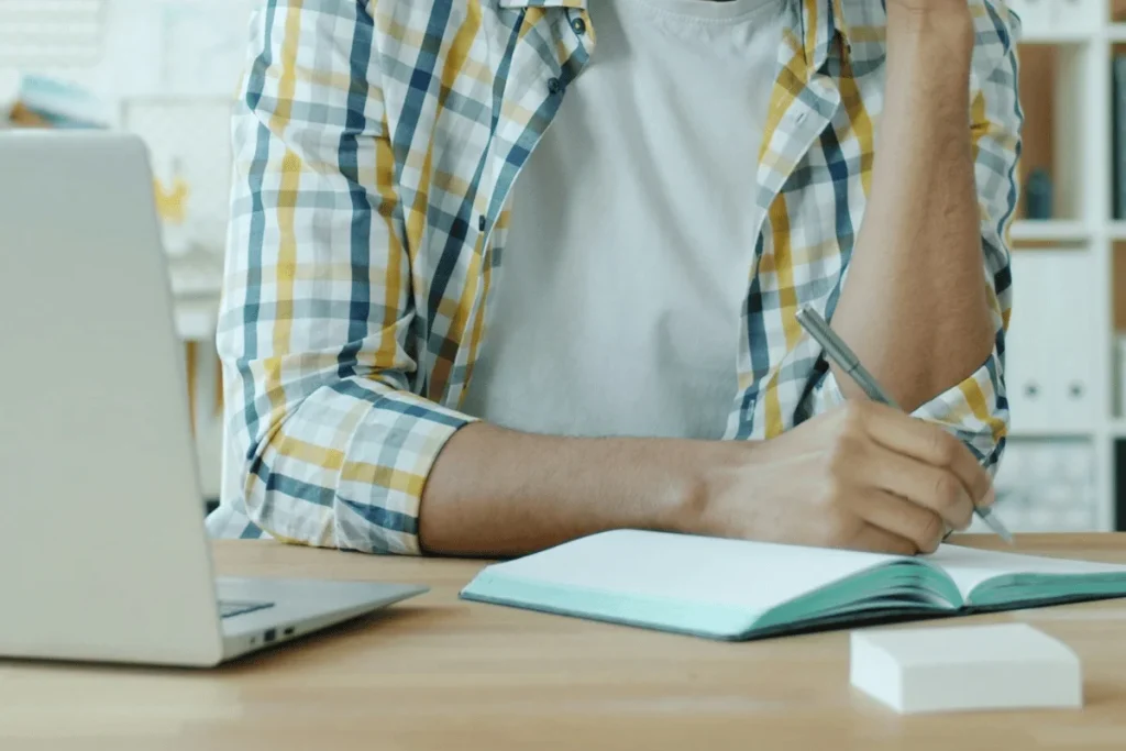 Person writing notes in a notebook beside a laptop.