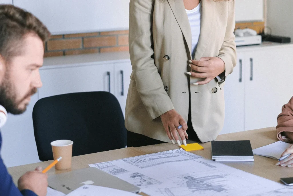 A person in a beige blazer gestures towards a table covered with blueprints and notes during a meeting.