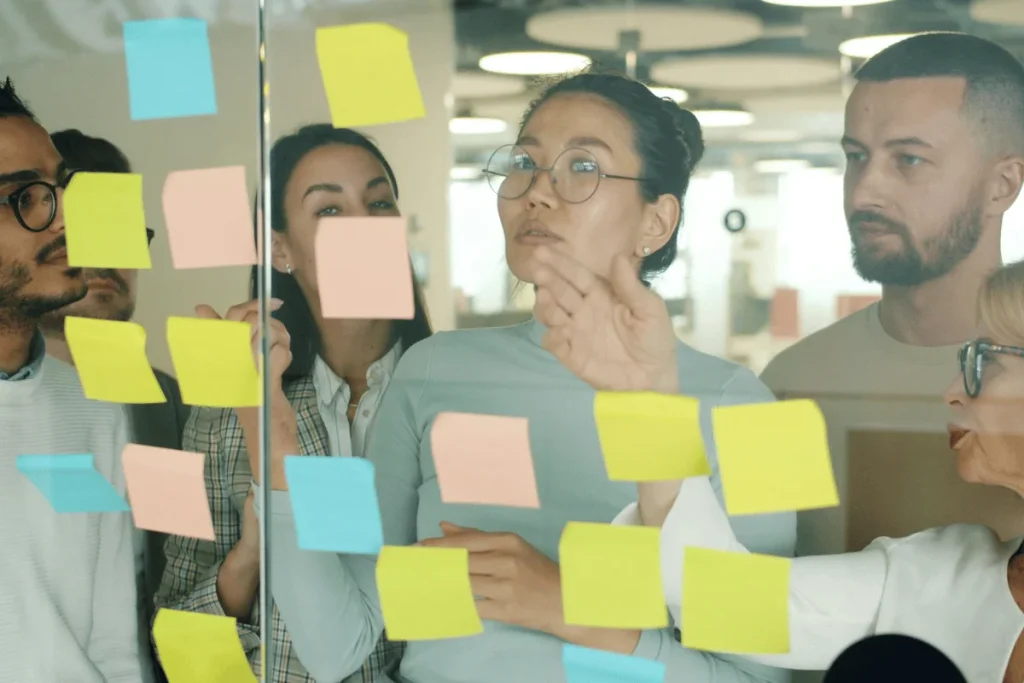 A diverse group of people in a meeting focuses on colorful sticky notes on a glass wall.