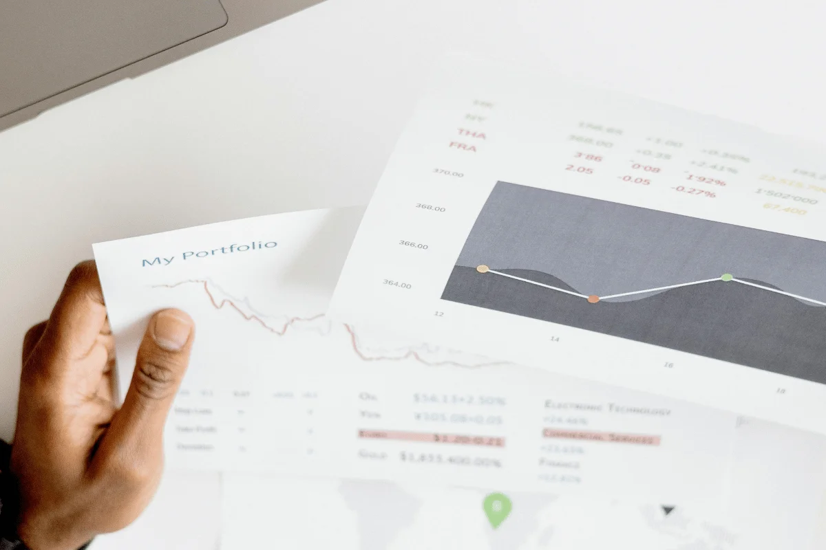 A person's hand holds financial documents showing a line graph and a portfolio report. 