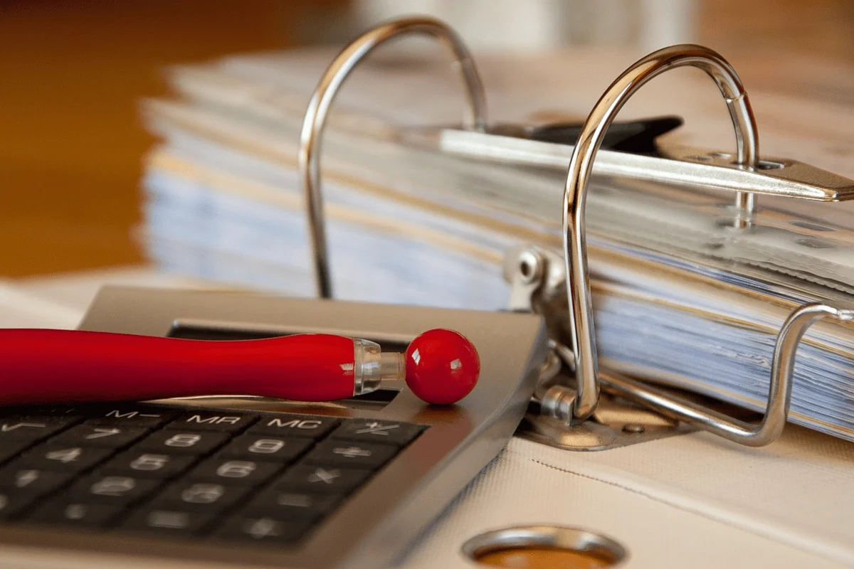 A close-up of a gray calculator with red buttons and a red pen resting on it, beside a thick open binder filled with paper, suggesting an office setting.