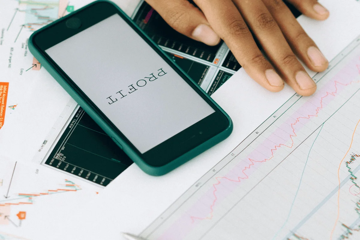 A hand rests on financial charts beside a smartphone displaying the word "PROFIT."