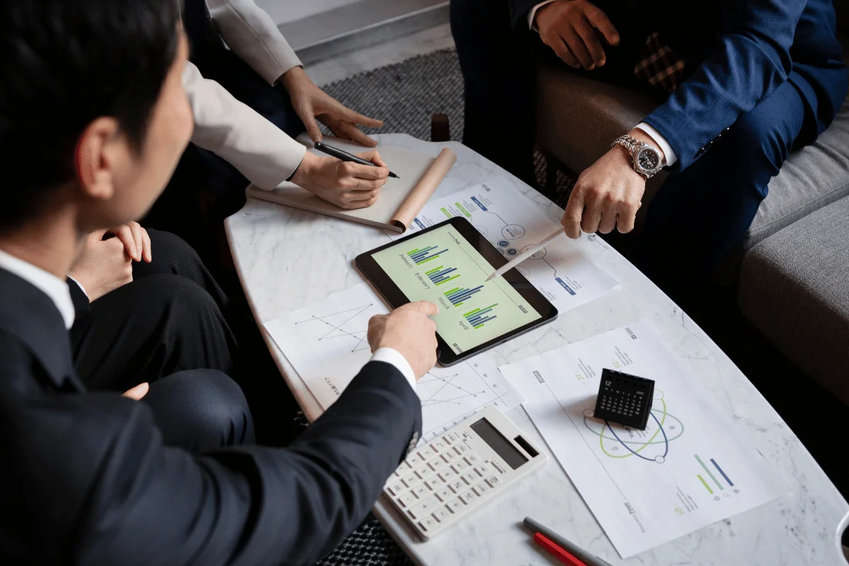 A business meeting with four people discussing data on a tablet, surrounded by papers, a calculator, and a desk calendar.
