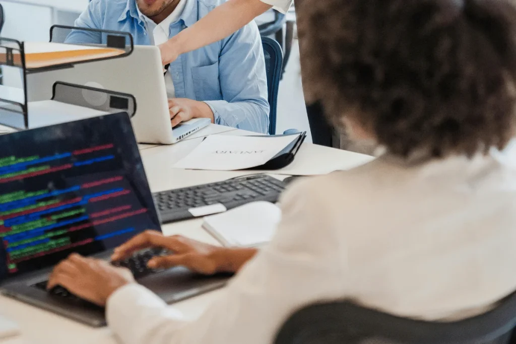 A person types code on a laptop, displaying colorful text. Another person in a blue shirt works at a desk nearby.