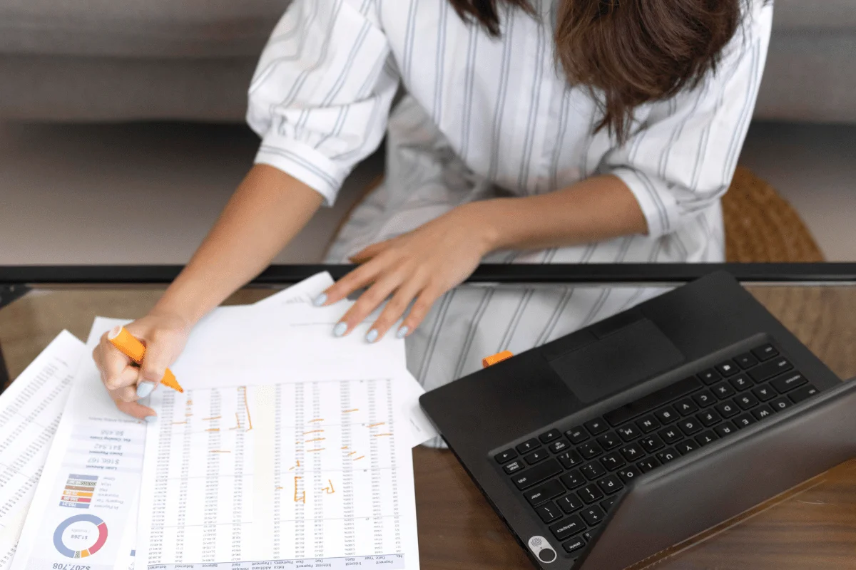A person in a striped shirt is reviewing charts and data with an orange pen, while seated at a glass table with a black laptop. 