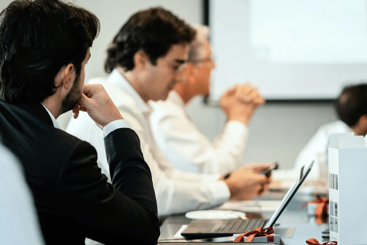 People in a conference room, dressed in formal attire, focused on a presentation. Laptops and notepads are on the table, suggesting a professional setting.