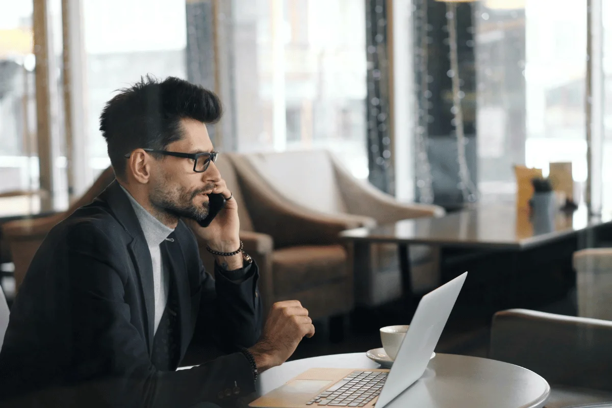 A man in a suit and glasses talks on the phone while using a laptop in a cozy cafe. 